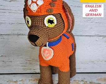 Op de afbeelding: Gehaakt knuffeldier in de vorm van een hond, die een oranje jumpsuit draagt met een witte voetafdruk en een blauwe halsband met een roze hartbedel. De hond draagt een oranje helm met een witte voetafdruk.
