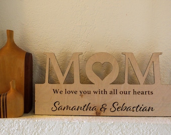 May include: Wooden sign with the word "MOM" in large cutout letters, a heart cutout in the centre, and the text "We love you with all our hearts Samantha & Sebastian" below.