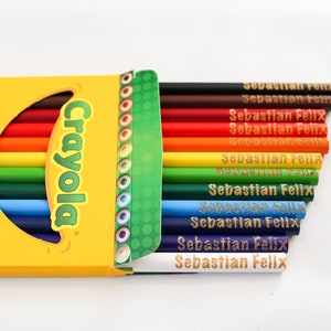May include: A yellow Crayola crayon box with a selection of coloured pencils. The box is open, revealing a rainbow of pencils, each labelled "Sebastian Felix." The box has text that says "Made with Forested Wood" and "Preferred by Teachers!"