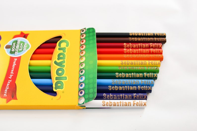 May include: A yellow Crayola crayon box with a selection of coloured pencils. The box is open, revealing a rainbow of pencils, each labelled "Sebastian Felix." The box has text that says "Made with Forested Wood" and "Preferred by Teachers!"