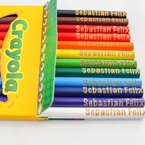 May include: A selection of Crayola coloured pencils in a range of hues, from red to purple. Each pencil is inscribed with the name "Sebastian Felix" in gold. The pencils are presented in a yellow box featuring the Crayola logo.