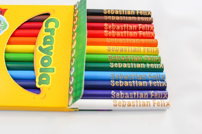 May include: A selection of Crayola coloured pencils in a range of hues, from red to purple. Each pencil is inscribed with the name "Sebastian Felix" in gold. The pencils are presented in a yellow box featuring the Crayola logo.