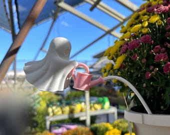 May include: A white ghost figurine holding a small pink watering can, attached to a white stake inserted into a potted plant. The ghost has a smooth, rounded head and a flowing, draped body shape, while the watering can features a handle.