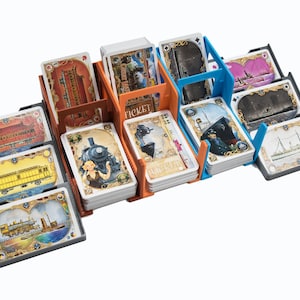 Op de afbeelding: Een spelorganizer voor het bordspel Ticket to Ride. De organizer is gemaakt van oranje en blauw plastic en heeft vakken voor verschillende soorten kaarten. De organizer wordt getoond met kaarten in elk vak.