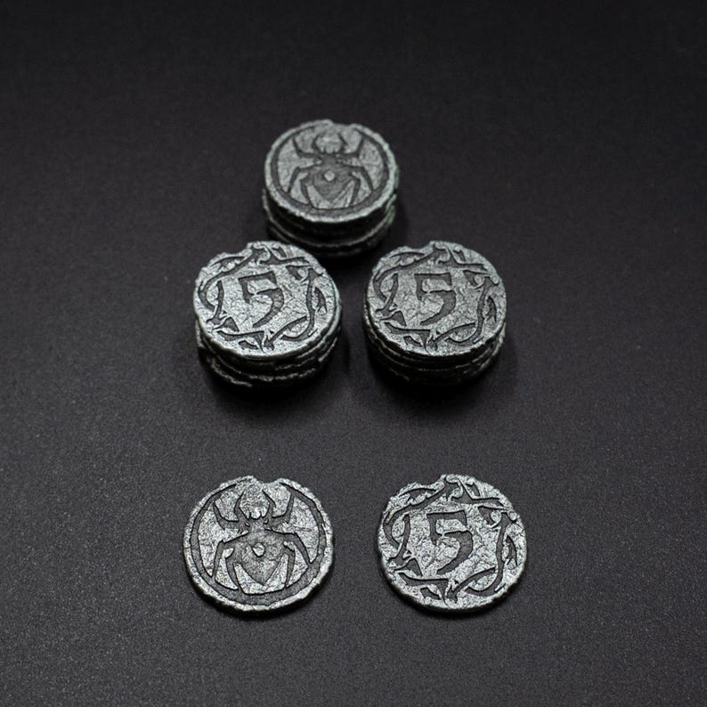 Peut inclure: Collection de jetons de jeu argent&eacute;s antiques. Les jetons pr&eacute;sentent un motif d'araign&eacute;e et le chiffre "5" avec des volutes d&eacute;coratives. Les jetons sont empil&eacute;s en petits tas et dispos&eacute;s sur une surface sombre.