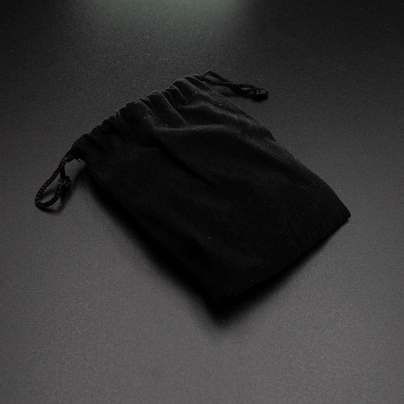 Peut inclure: Un sac en velours noir avec cordon de serrage, id&eacute;al pour les bijoux ou les petits cadeaux. Le sac est ferm&eacute; par un cordon noir et pos&eacute; sur un fond sombre.