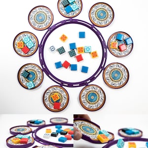 Può includere: Un tabellone da gioco circolare viola con otto porta tessere colorati. I porta tessere hanno un motivo decorativo e sono disposti in cerchio. Ci sono piccole tessere colorate sparse intorno al tabellone.