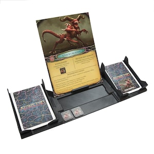 Puede incluir: Un organizador de juegos de cartas negro con una tarjeta titulada "GNIOWORODNY" y el texto "WYZWOLENIE" y "TRUDNIEJSZY WARIANT ZASADY" visible. El organizador tiene dos ranuras para barajas de cartas y dos ranuras para dados.
