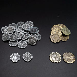 Könnte beinhalten: Eine Sammlung von Spielmarken aus Metall in Silber- und Bronzetönen. Die Marken weisen verschiedene Designs auf, darunter die Zahl "1", ein stilisiertes "T" und der Text "Concorde Espadon". Die Marken sind auf einer dunklen Oberfläche angeordnet.