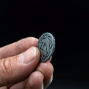 Peut inclure: Une petite pi&egrave;ce de monnaie gris fonc&eacute; avec un motif en relief est tenue entre les doigts sur un fond noir. La surface de la pi&egrave;ce pr&eacute;sente un motif complexe, comprenant peut-&ecirc;tre du texte ou des symboles. Les bords de la pi&egrave;ce sont l&eacute;g&egrave;rement irr&eacute;guliers.