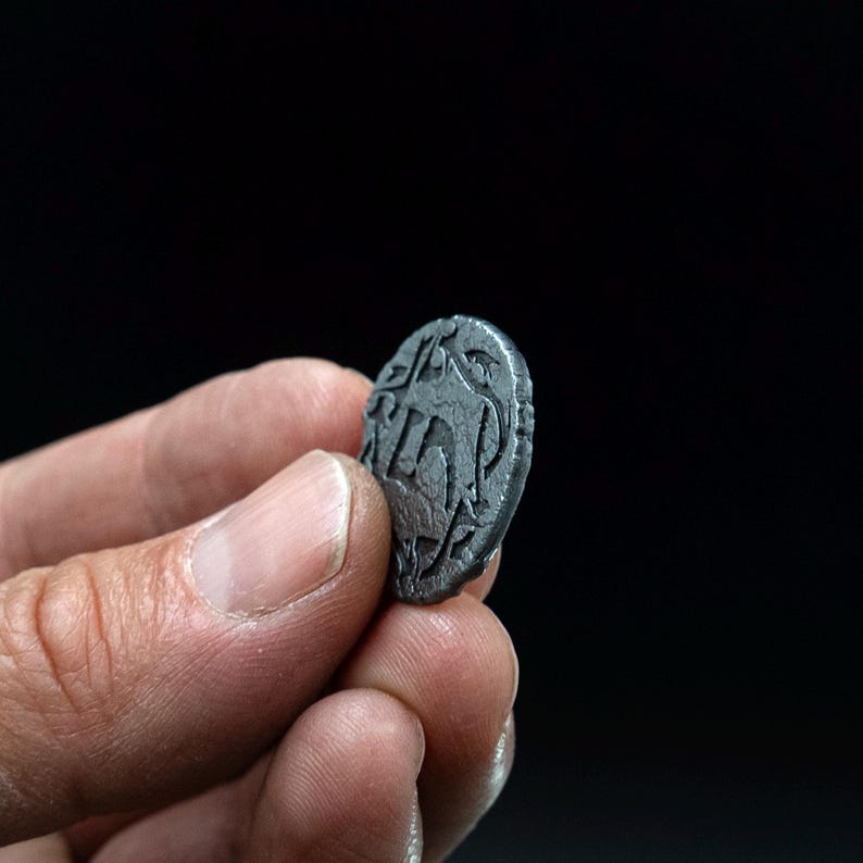 Peut inclure: Une petite pi&egrave;ce de monnaie gris fonc&eacute; avec un motif en relief est tenue entre les doigts sur un fond noir. La surface de la pi&egrave;ce pr&eacute;sente un motif complexe, comprenant peut-&ecirc;tre du texte ou des symboles. Les bords de la pi&egrave;ce sont l&eacute;g&egrave;rement irr&eacute;guliers.