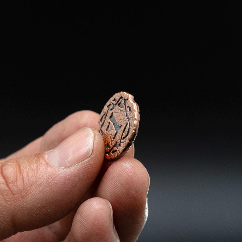 Peut inclure: Une petite pi&egrave;ce de monnaie de couleur cuivr&eacute;e avec un motif en relief. La pi&egrave;ce est tenue entre les doigts sur un fond sombre. Le motif semble &ecirc;tre une figure ou un symbole stylis&eacute; dans un cadre circulaire.