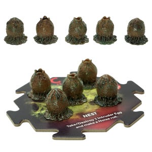 Può includere: Un set di pezzi di gioco a forma di uovo marrone strutturato con una base verde. Sono esposte sei uova, cinque separate e una su un tabellone di gioco. Il tabellone riporta la parola "NEST" e le istruzioni di gioco.