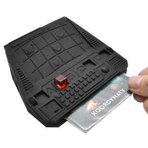 May include: A black plastic game console with a red button and a card that says "KOORDYNATY" in the slot. The console has a grid of squares on the top and a keyboard on the bottom.