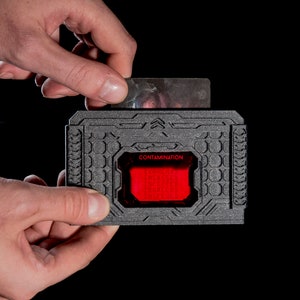 Puede incluir: Un porta tarjetas negro con una luz roja y la palabra "CONTAMINATION" impresa en él. El porta tarjetas tiene una superficie texturizada y una ranura para insertar una tarjeta.