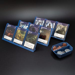 May include: A blue plastic gaming card organiser with multiple slots holding various illustrated fantasy cards, accompanied by a separate blue hexagonal tray containing a collection of small metallic coins with embossed circular designs.