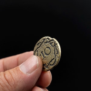 Peut inclure: Pi&egrave;ce de monnaie en bronze vieilli avec le chiffre 10 au centre, entour&eacute;e d'un motif ornemental. La pi&egrave;ce est tenue entre les doigts sur un fond noir. La surface de la pi&egrave;ce montre des signes d'usure, sugg&eacute;rant l'&acirc;ge et l'utilisation.