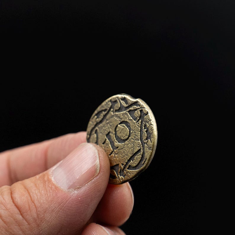 Peut inclure: Pi&egrave;ce de monnaie en bronze vieilli avec le chiffre 10 au centre, entour&eacute;e d'un motif ornemental. La pi&egrave;ce est tenue entre les doigts sur un fond noir. La surface de la pi&egrave;ce montre des signes d'usure, sugg&eacute;rant l'&acirc;ge et l'utilisation.