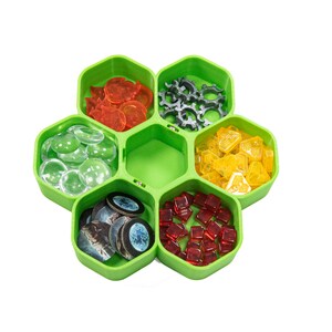 Könnte beinhalten: Ein grünes sechseckiges Tablett mit sechs Fächern, die jeweils mit verschiedenen farbigen Spielsteinen gefüllt sind. Die Fächer enthalten klare Glasstücke, rote Glassteine, schwarze und weiße runde Stücke, gelbe Plastikstücke, graue Metallräder und orangefarbene Plastikstücke.