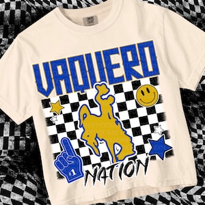 May include: A beige t-shirt featuring a blue and gold graphic of a cowboy riding a bucking horse. The graphic is set against a black and white chequered background. The text "Vaquero Nation" is printed below the graphic.