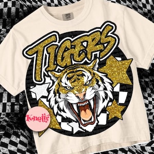 May include: A beige t-shirt with a black and gold graphic of a tiger with the word "TIGERS" in gold glitter letters. The graphic is surrounded by gold glitter stars.