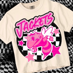 May include: A pink and black graphic tee with the word "Jackets" in a graffiti style font. A pink bee with wings is in the center of the design, surrounded by pink stars and a black and white checkered pattern.