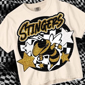 May include: A beige t-shirt with a black and white chequered pattern and a gold glitter design. The design features a bee with wings, the word "Stingers" in gold glitter, and stars.