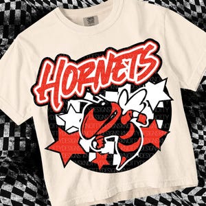 Peut inclure: Un t-shirt beige avec un graphique noir et rouge d'un frelon avec le mot "HORNETS" en lettres rouges et noires.