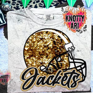 May include: A white t-shirt with a distressed design featuring a football helmet filled with gold sequins. The word "Jackets" is written in a script font below the helmet. The shirt has a crew neck and short sleeves.
