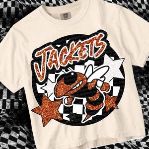 May include: Cream-colored t-shirt featuring a cartoon bee mascot with the word "JACKETS" in glittery orange letters. The bee is orange and black, set against a black circle with white stars and a checkered background.