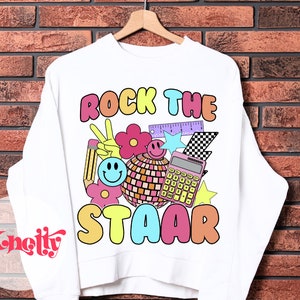Può includere: Felpa bianca con un design grafico colorato che dice "Rock the STAAR". La grafica include un segno di pace, una matita, una calcolatrice, un righello, una palla da discoteca, una stella e fiori.
