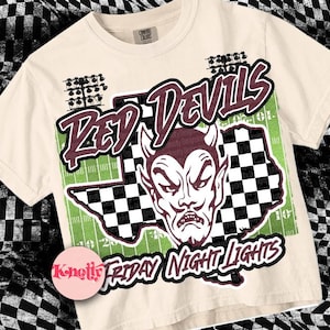 May include: A beige t-shirt with a graphic print of a red devil with horns and a checkered flag design. The text "Red Devils" and "Friday Night Lights" is printed on the shirt.