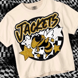May include: A beige t-shirt with a black and gold graphic design. The graphic features the word "Jackets" in a graffiti style font, a bee with wings, and stars.