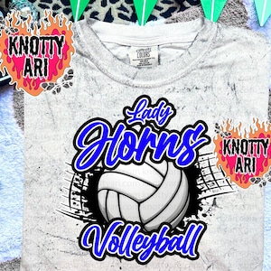 May include: A light gray t-shirt with a volleyball graphic. The graphic features the words "Lady Horns Volleyball" in blue and white. The shirt has a crew neck and short sleeves.