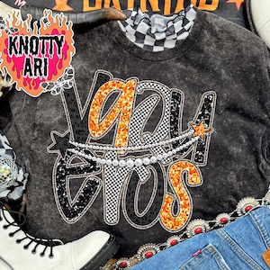 Op de afbeelding: Een donkergrijs sweatshirt met de tekst "Vegas Eros" in oranje glitter, zwarte luipaardprint en geblokte patronen. Een parelketting en steraccenten sieren het ontwerp. De trui heeft een geblokte kraag.