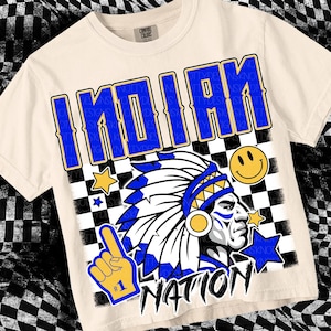 May include: A beige t-shirt with a blue and yellow graphic design. The graphic features a Native American headdress, a smiley face, and the text "INDIAN NATION".
