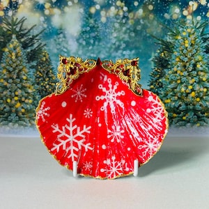 May include: A red scallop shell painted with white snowflakes and white dots, featuring a gold-toned filigree metal ornament at the top edge adorned with small red crystals. The shell has a gold-painted rim and is displayed on a small white stand.