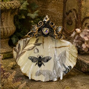 May include: A decorative scallop shell dish featuring a black and white bee illustration, botanical prints, and gold-painted edges. The top is adorned with an ornate black and gold metal crest featuring a central iridescent heart-shaped crystal.