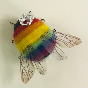 May include: A handmade bee-shaped brooch with a rainbow-colored body and clear wings. The bee's body is crafted from felted wool in vibrant colors, and it is topped with a small silver crown. The wings are translucent, adding a delicate touch to the design.