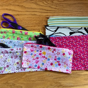 May include: A stack of eight zippered pouches in various floral and geometric patterns. The pouches are made of fabric and have a zipper closure. Some of the pouches have a white background with colorful flowers, while others have a solid color background with a geometric pattern. The pouches are arranged in a stack, with the top pouch being a green floral pattern.