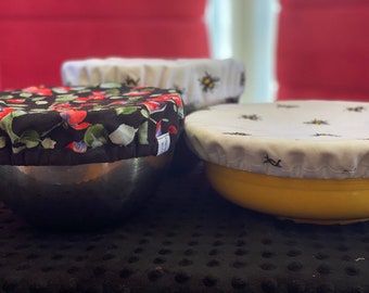 May include: Three reusable bowl covers. One is black with a floral pattern, one is white with a bee pattern, and one is white with a bee pattern on a yellow bowl.