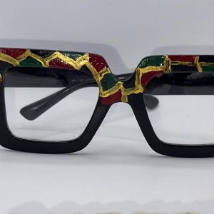 Puede incluir: Gafas cuadradas negras con lentes transparentes. La parte superior de las monturas está decorada con un patrón rojo, verde y dorado. Las gafas tienen un puente y patillas negros.