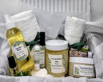 May include: A gift box filled with bath and body products, including a body butter, body polish, bath salts, a natural fruit inhaler, and a bottle of body oil. The box is lined with white tissue paper and has a green ribbon.