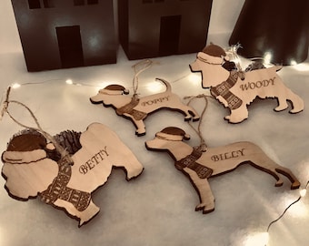 May include: Four wooden dog ornaments with the names "Betty", "Poppy", "Woody", and "Billy" engraved on them. Each ornament has a Santa hat and a scarf. The ornaments are on a white surface with white Christmas lights.