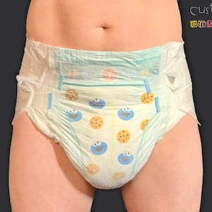 Puede incluir: Un pañal de color claro con un estampado de personajes de dibujos animados azules y galletas. El pañal tiene pestañas azules y está diseñado para la comodidad y la absorción. El texto "customZ diapers" es visible en la esquina superior derecha.
