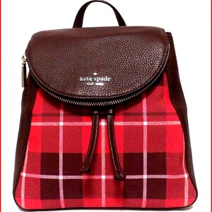 May include: A red plaid backpack with a brown leather flap and trim. The backpack has a drawstring closure and a top handle. The brand name "kate spade NEW YORK" is visible on the flap.