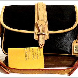 May include: Black handbag with a flap closure, tan trim, and a matching adjustable shoulder strap. The bag features a tan leather strap with a metal buckle and a small tag with the brand name. A yellow business card is visible.