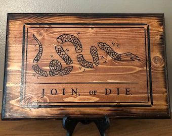 May include: Wooden sign with a dark brown border and a lighter brown centre. The sign features a segmented snake illustration with the words "JOIN, or DIE" in black lettering. The sign is displayed on a black stand.