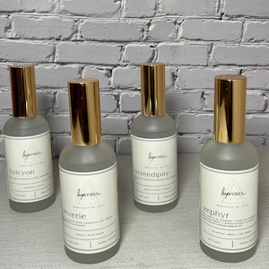 May include: Four glass bottles with gold caps and white labels. The labels have the text "luxe VIBES" and the names of different scents: "halcyon", "reverie", "serendipity", and "zephyr".