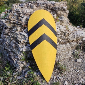 May include: A yellow shield with two black chevron stripes. The shield is mounted against a weathered rock formation, with green foliage in the background. The shield has a pointed bottom.